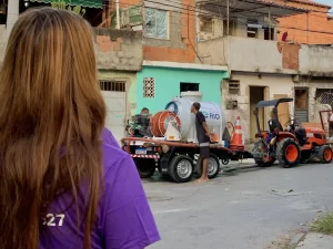 Uma pessoa de cabelo castanho claro e camiseta roxa está de costas, observando uma rua residencial com casas simples. Na rua, há um caminhão pequeno com equipamento de serviço e um trator laranja ao lado, indicando possíveis atividades de manutenção ou serviços públicos.